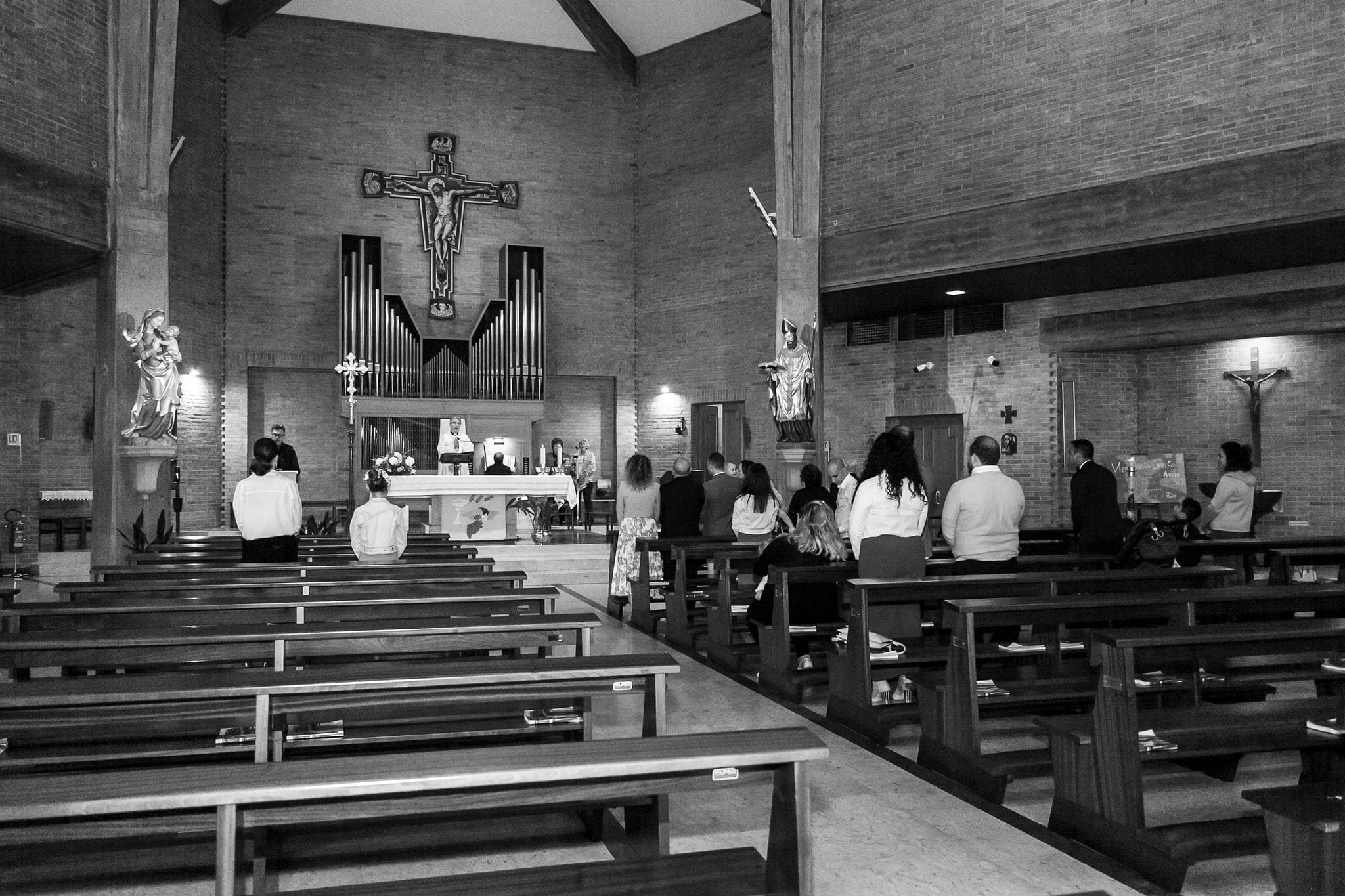 In bianco e nero, interno di una chiesa moderna con muro di mattoni, crocifisso, organo e invitati seduti tra le panche