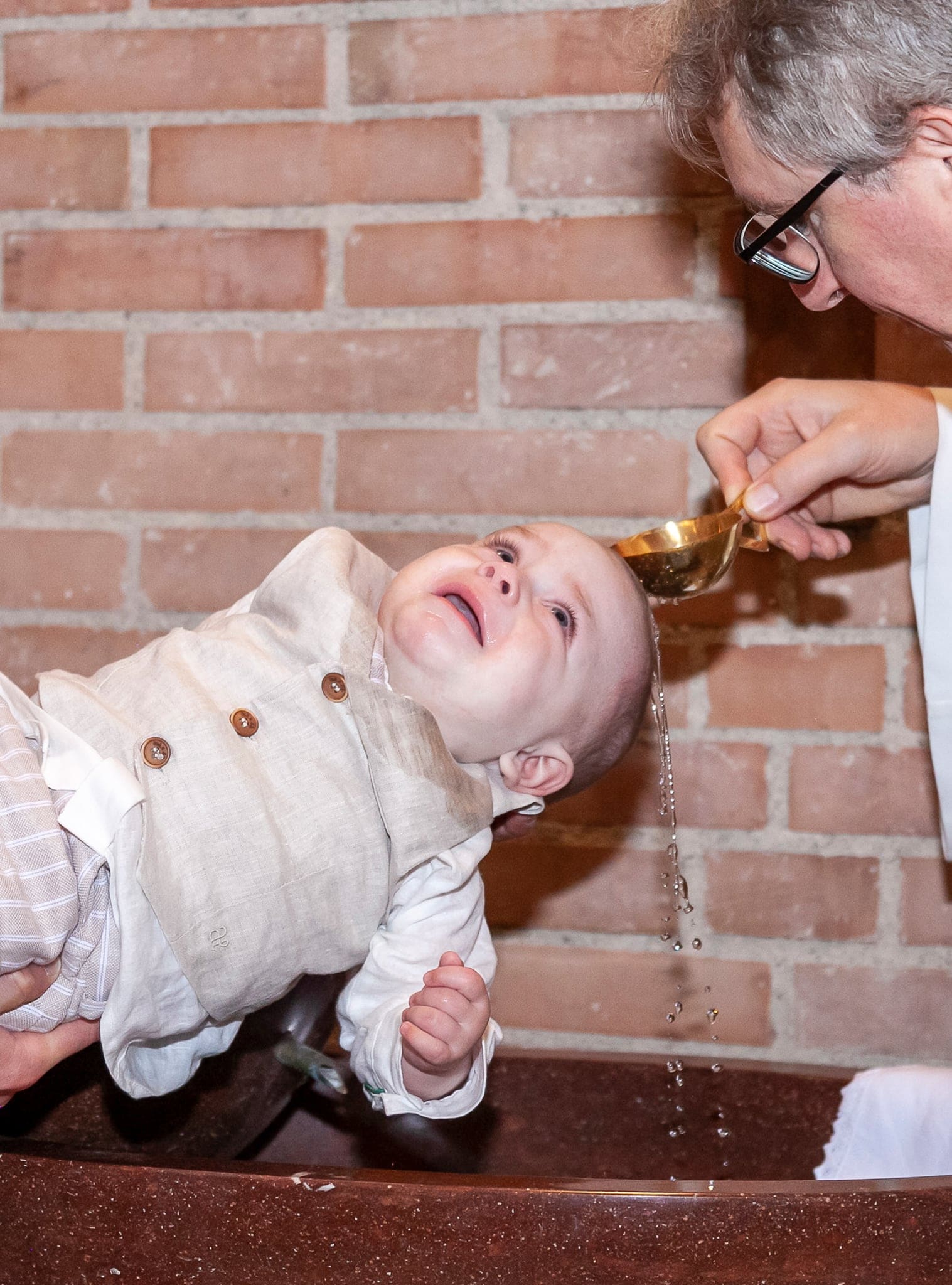 Il piccolo, con il gilet di lino, ride con la testa all'indietro mentre il sacerdote versa l'acqua dal cucchiaio sul fonte