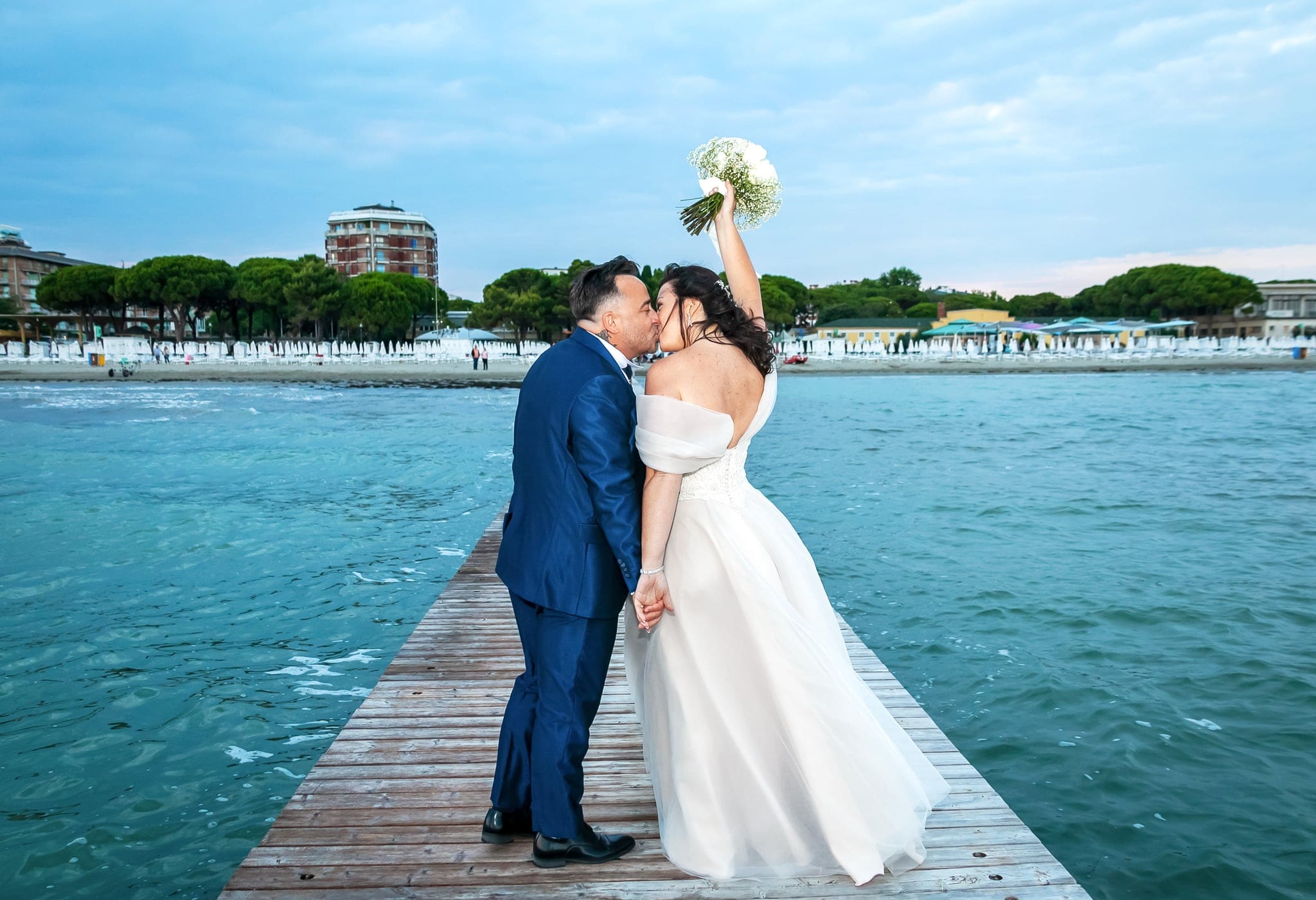 Sposi che si baciano sul molo con il bouquet alzato, il mare turchese e la costa di Grado sullo sfondo