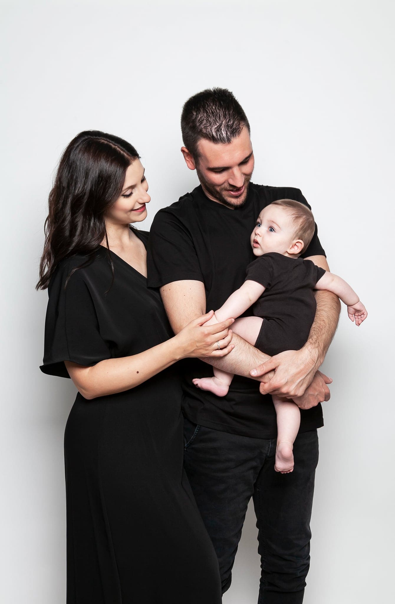 Mamma e papà in nero sorridono al loro bebè in braccio al papà, su fondo chiaro in studio.