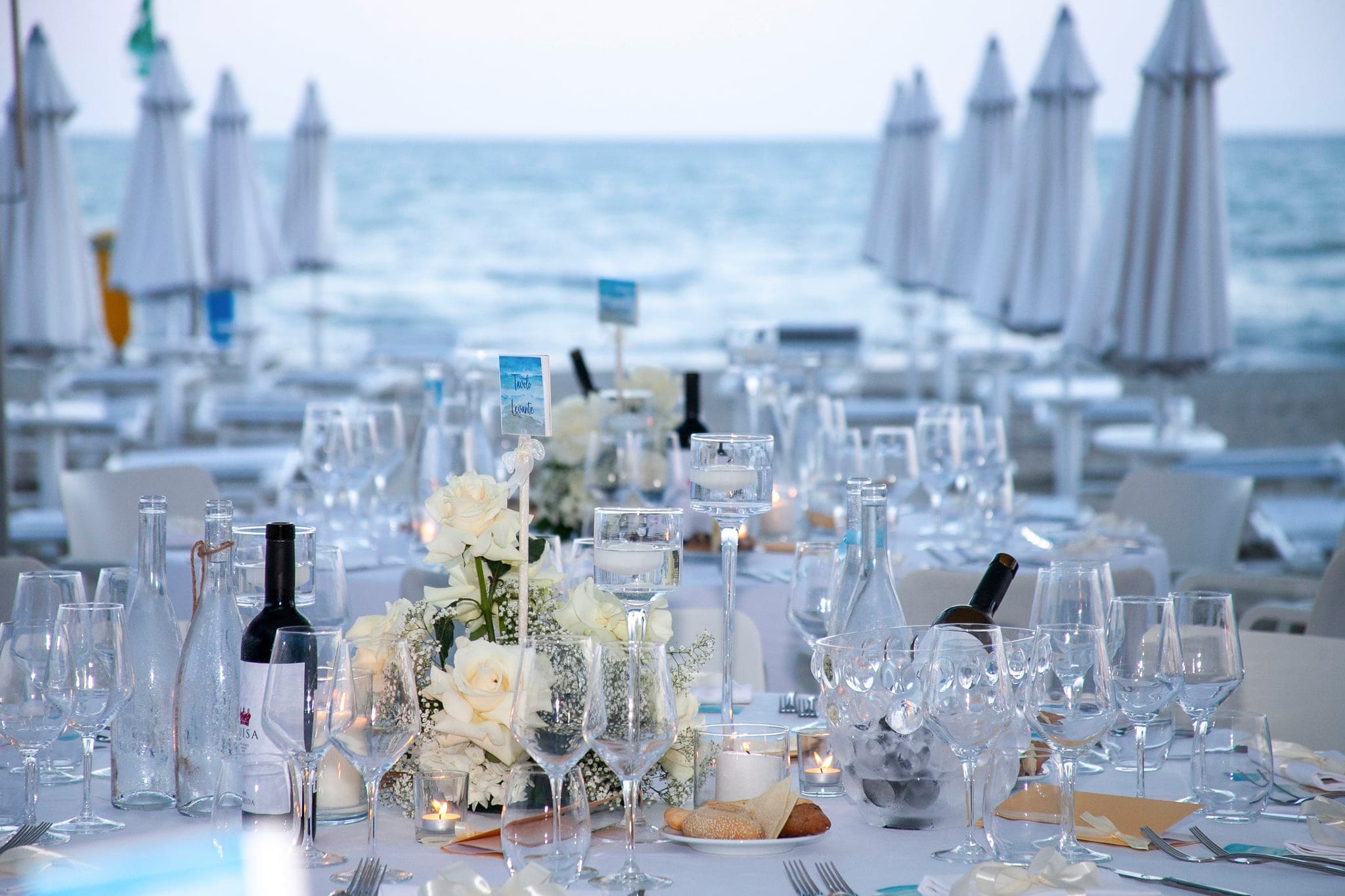 Tavolo apparecchiato con rose bianche e bicchieri sulla spiaggia, con ombrelloni chiusi e mare sullo sfondo, matrimonio a Grado