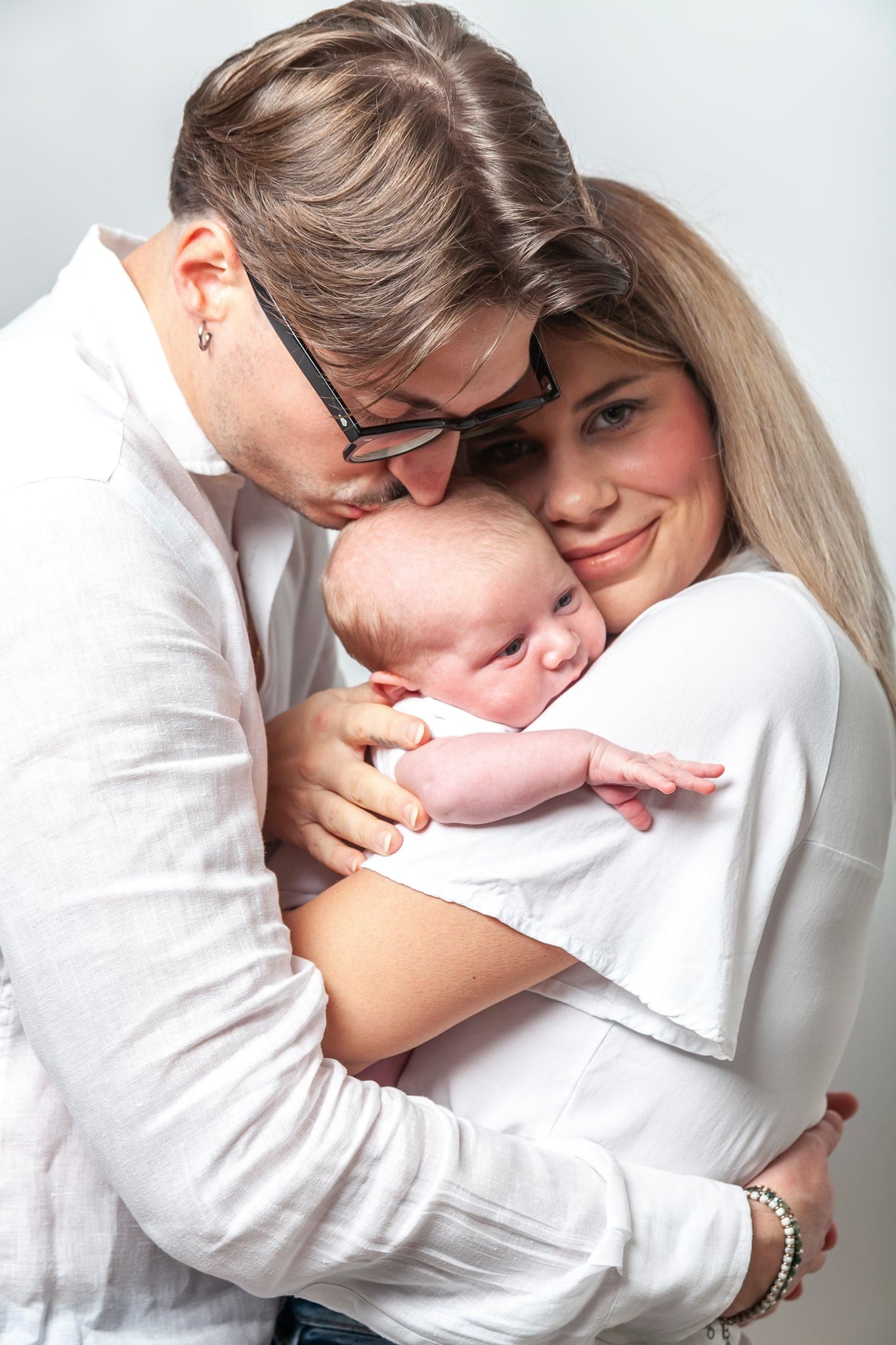 Papà bacia la fronte della mamma che stringe a sé il bebè, entrambi in abiti bianchi, in un abbraccio di famiglia.