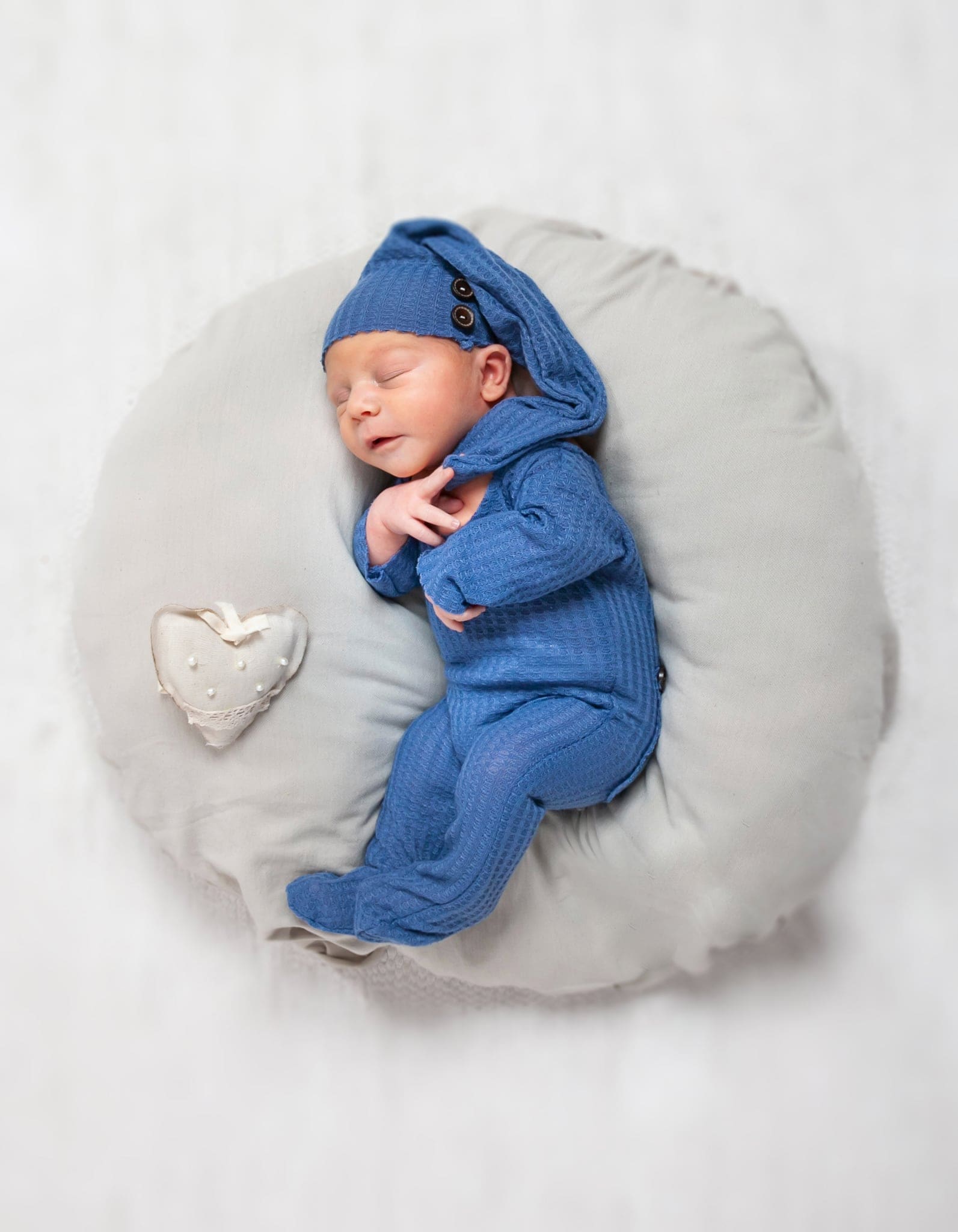 Neonato che dorme in tutina blu con cuffia sopra un cuscino tondo grigio, con un piccolo cuore decorativo, su fondo bianco.