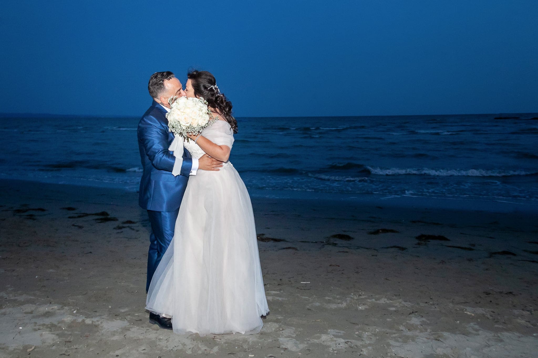 Sposi che si abbracciano e si baciano sulla riva al crepuscolo, con il mare blu alle spalle, matrimonio a Grado