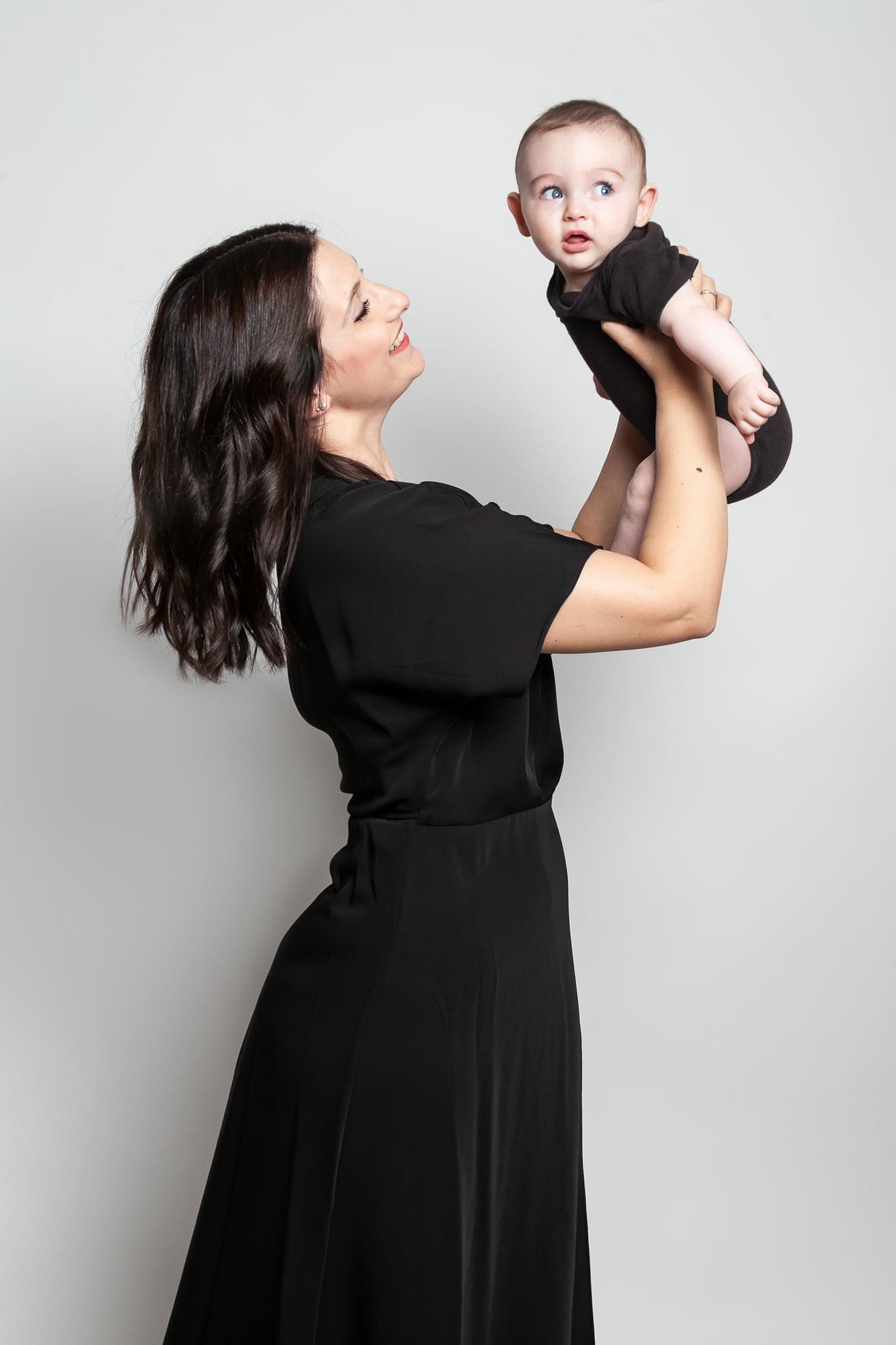Mamma in abito nero lunga che solleva in alto il suo bebè con un body scuro, sorridendogli, su fondo grigio chiaro.