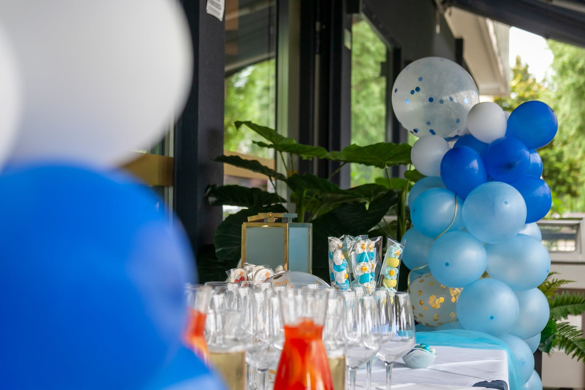 Colonna di palloncini azzurri, bianchi e coriandoli accanto a un tavolo apparecchiato con bicchieri, sfocati in primo piano
