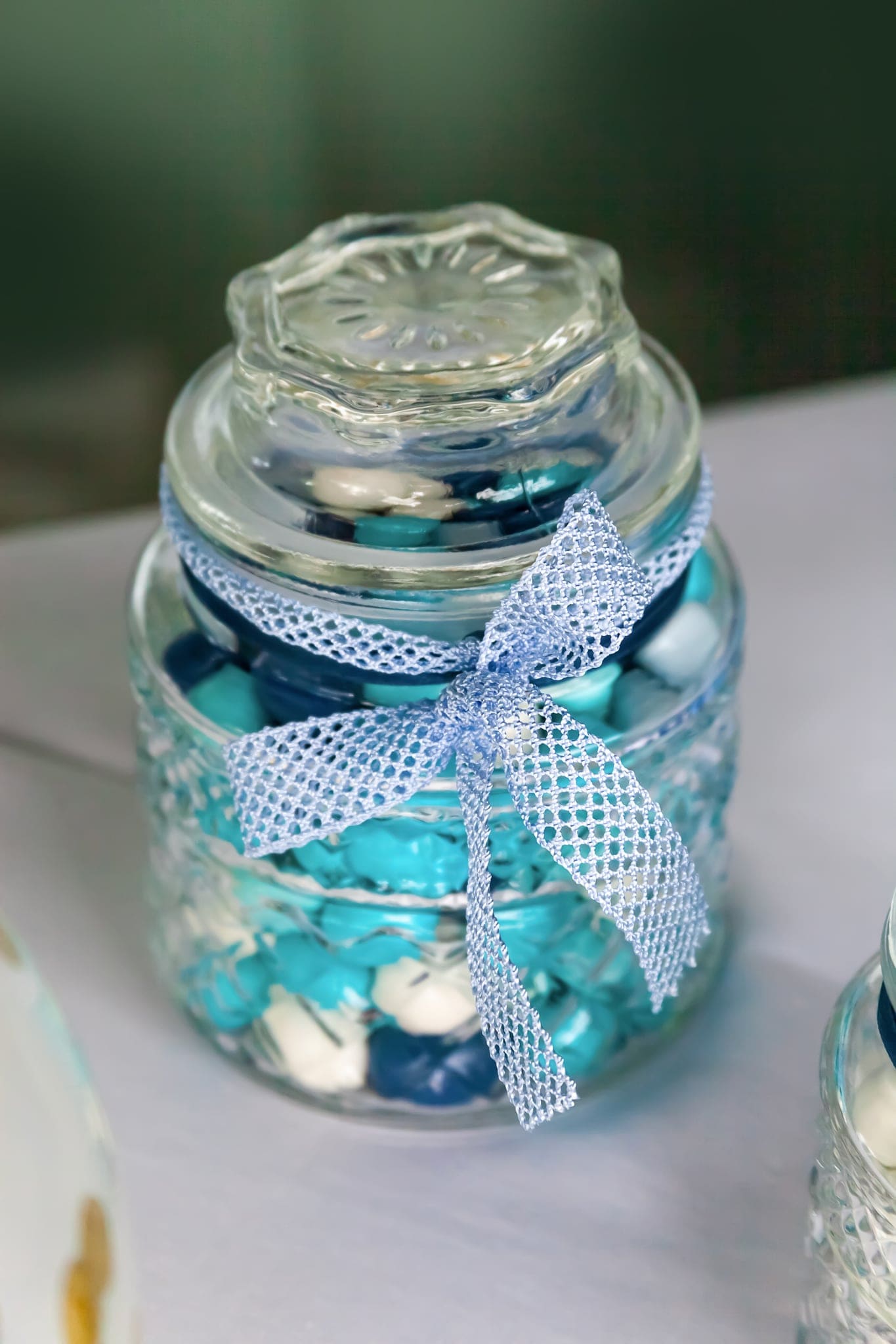 Barattolo di vetro con confetti azzurri e bianchi chiuso da un fiocco di rete azzurra, su una tovaglia chiara