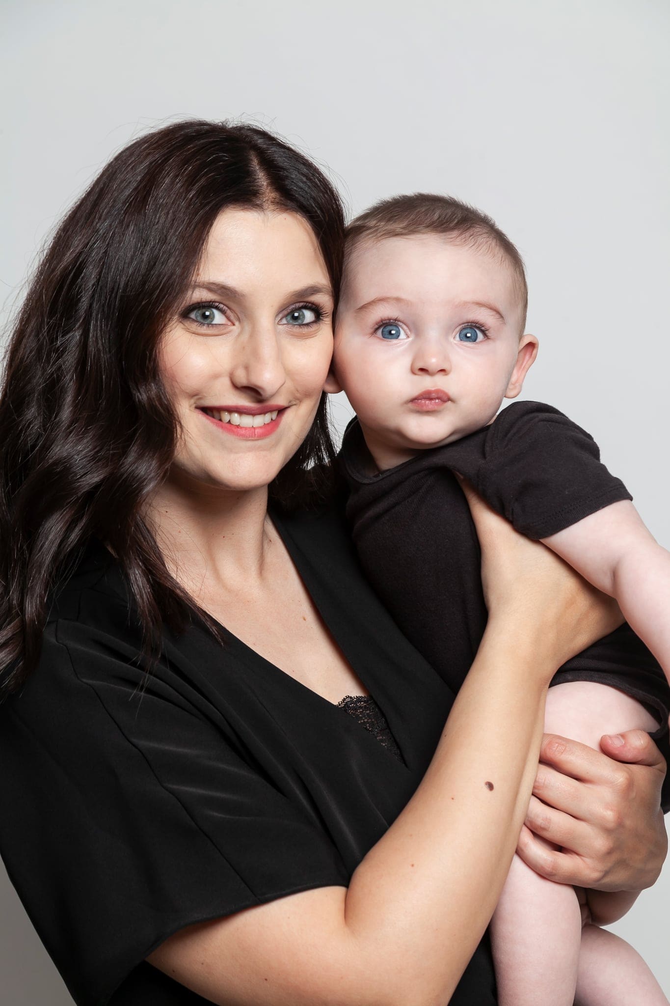 Ritratto di una mamma sorridente con il suo bebè in braccio che guarda in camera con occhi chiari, su fondo grigio.