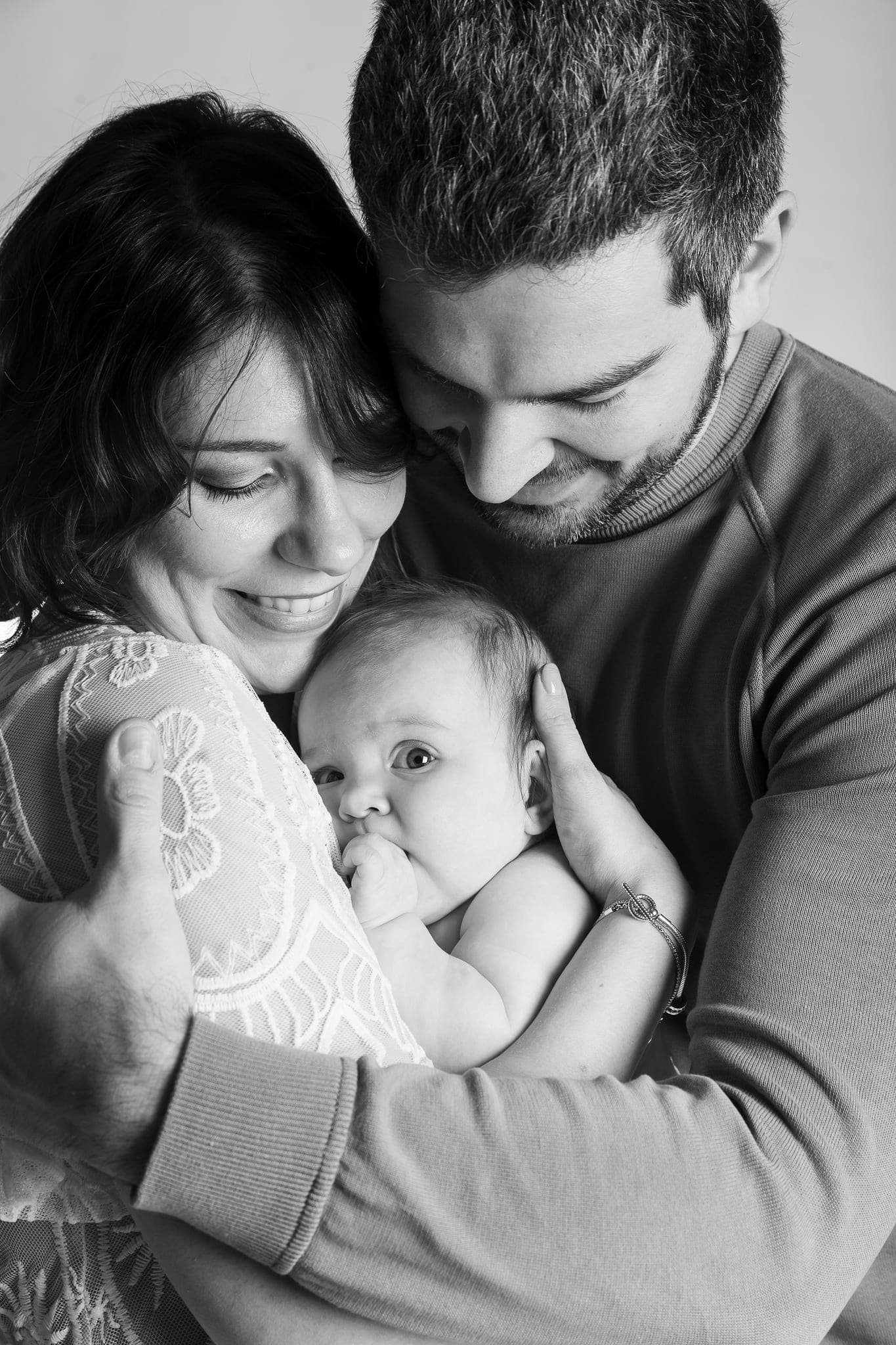 Mamma e papà che abbracciano il bebè, che si succhia il pugnetto, in uno scatto in bianco e nero ravvicinato.