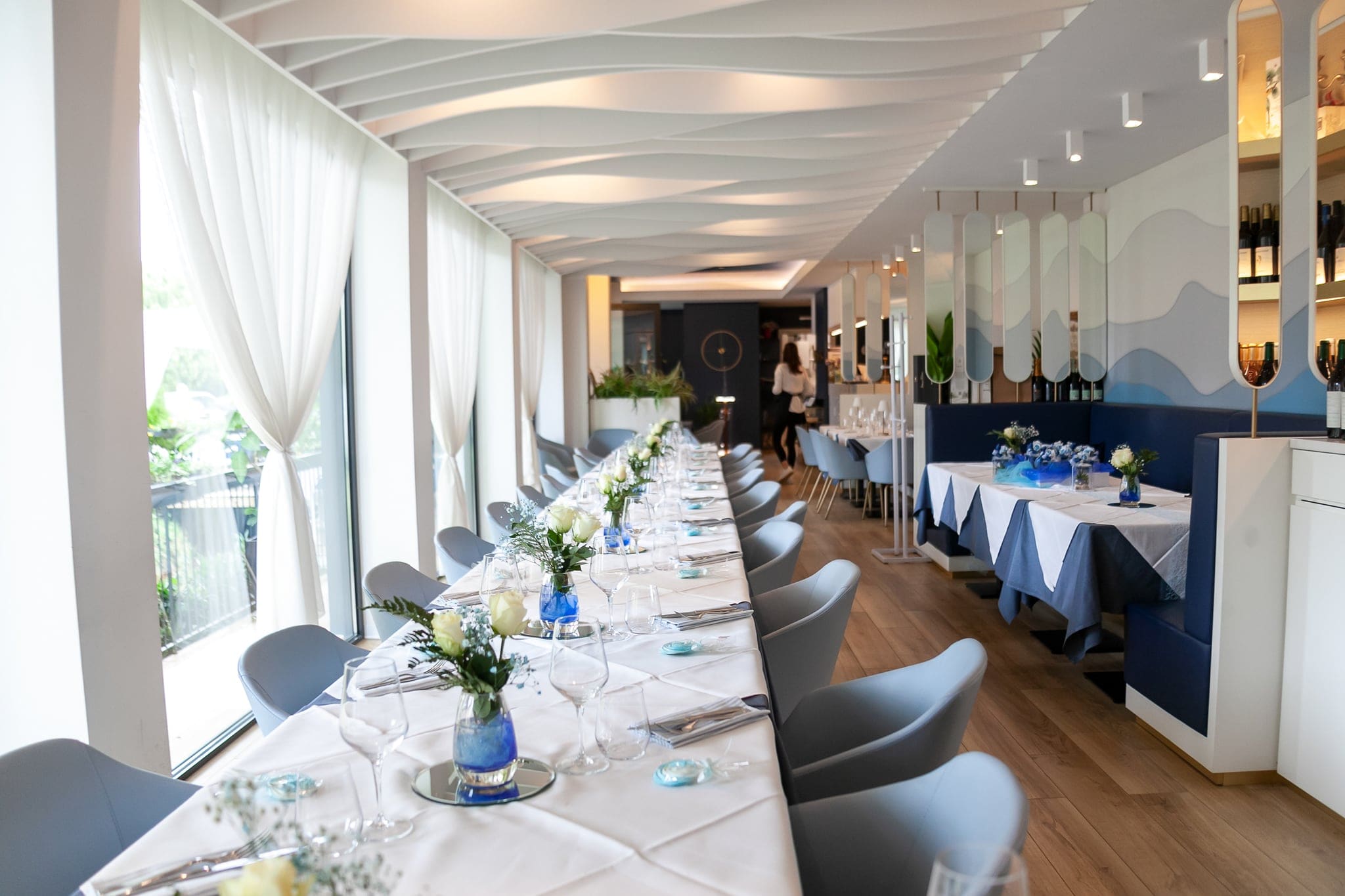 Sala del ristorante con la lunga tavolata apparecchiata in bianco e azzurro, sedie grigie e tende chiare alle ampie finestre