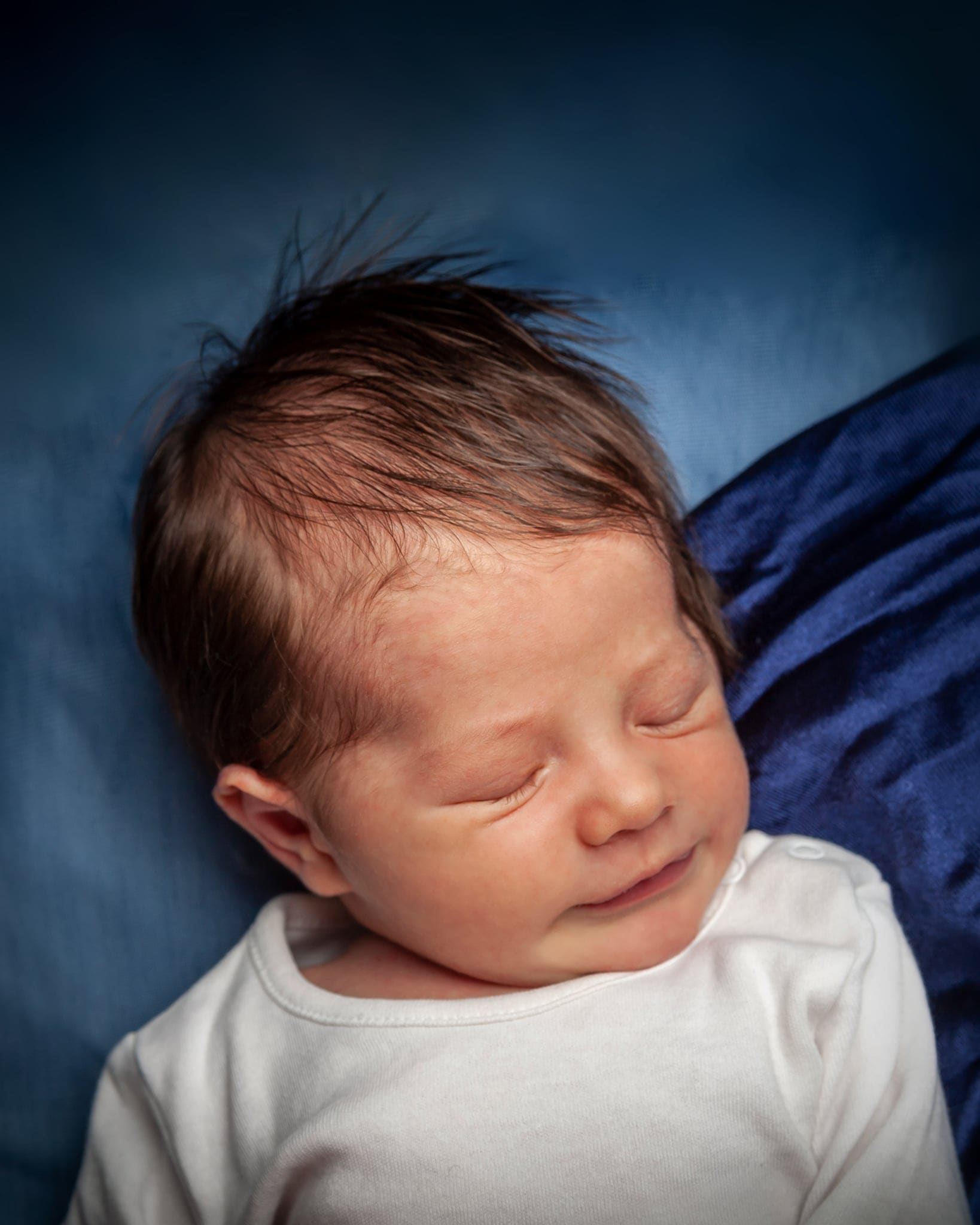 Ritratto di un neonato che dorme con capelli scuri e body bianco, su un telo blu.