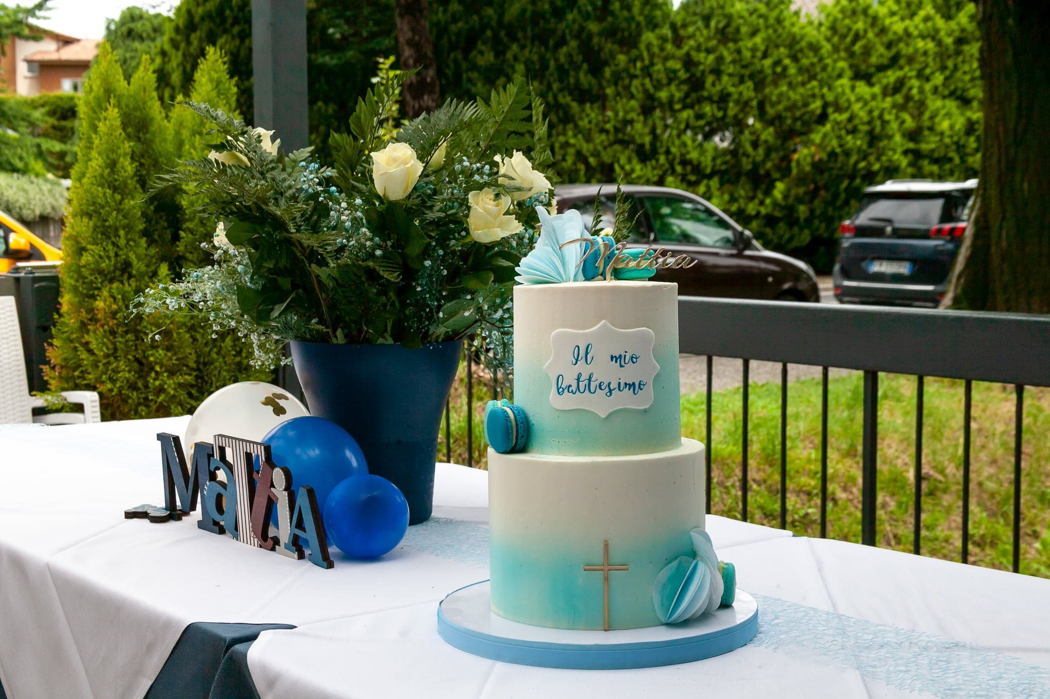 Torta a due piani sfumata di azzurro con macaron e la scritta Il mio battesimo, sul tavolo bianco accanto a fiori e palloncini
