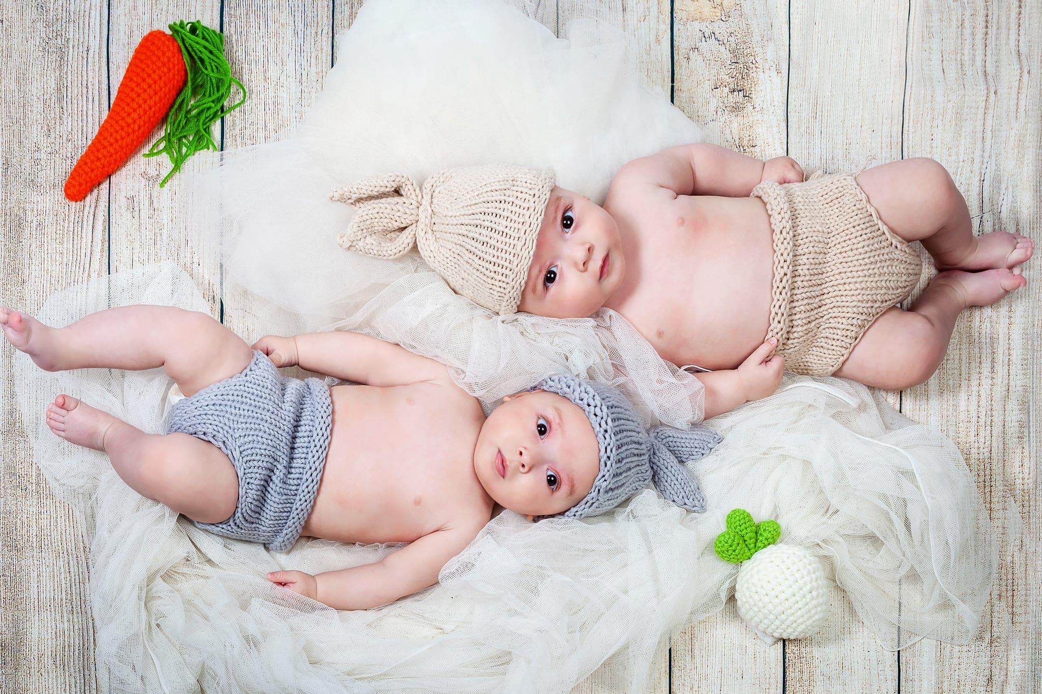 Due bebè sdraiati su un morbido tulle bianco, con cuffie e mutandine di lana beige e grigia, una carota e una rapa all'uncinetto.