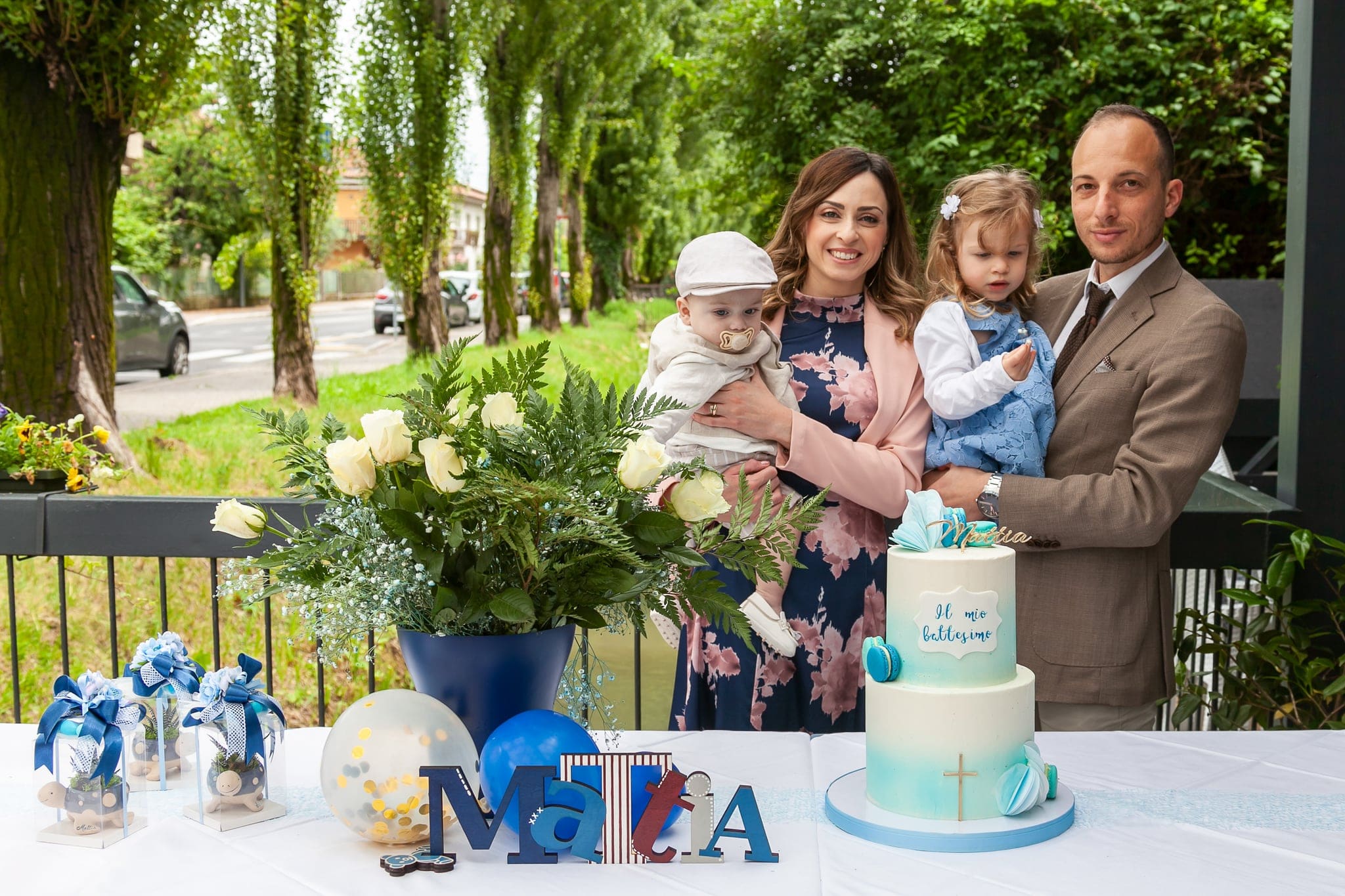 Mamma, papà, sorellina e il piccolo posano sorridendo dietro la torta azzurra e i palloncini, sulla terrazza in festa