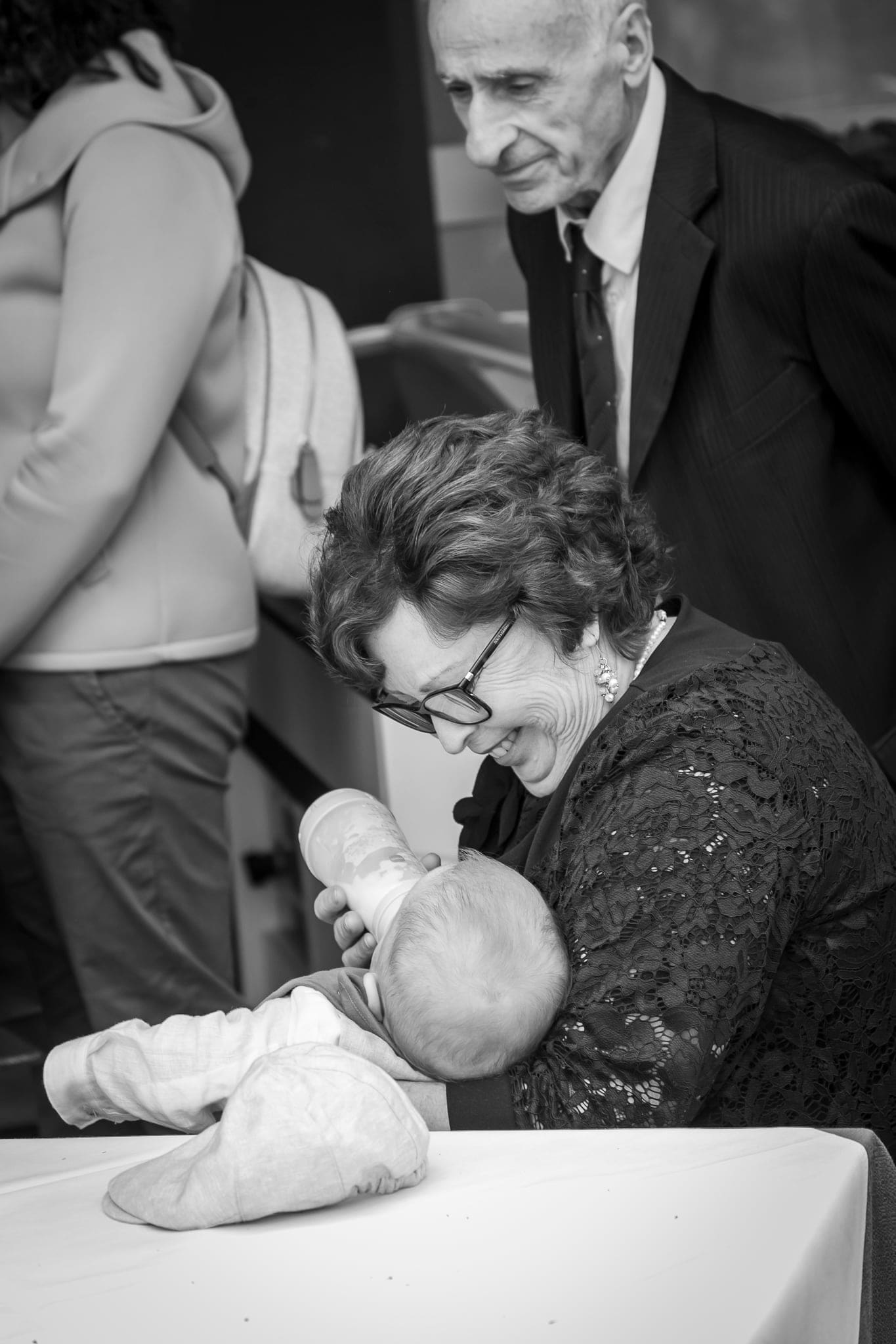 In bianco e nero, una nonna con gli occhiali si china sorridendo sul neonato, mentre un signore anziano osserva da dietro