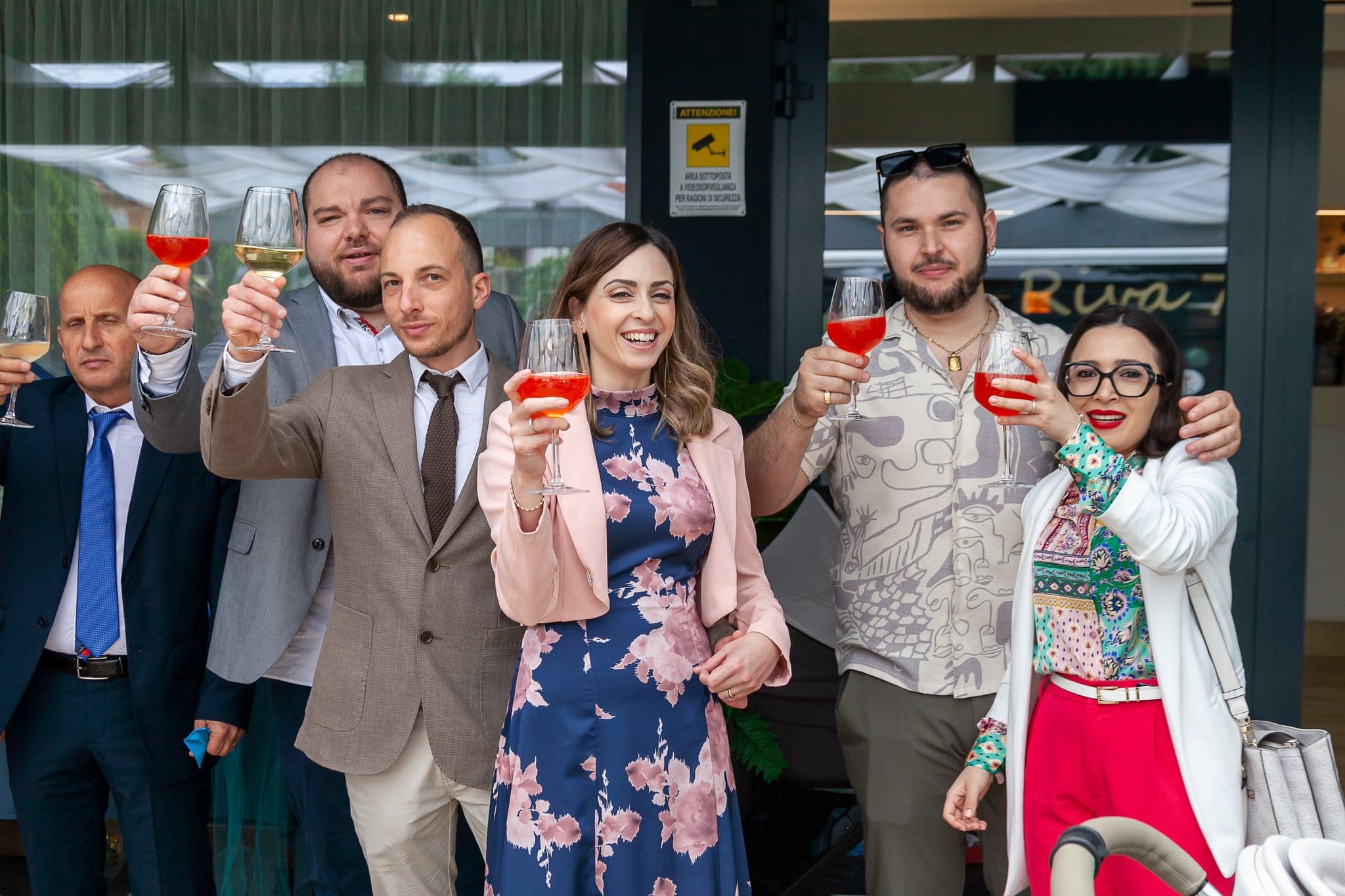 Un gruppo di invitati alza i calici in un brindisi sorridendo, davanti all'ingresso del locale dove si tiene la festa