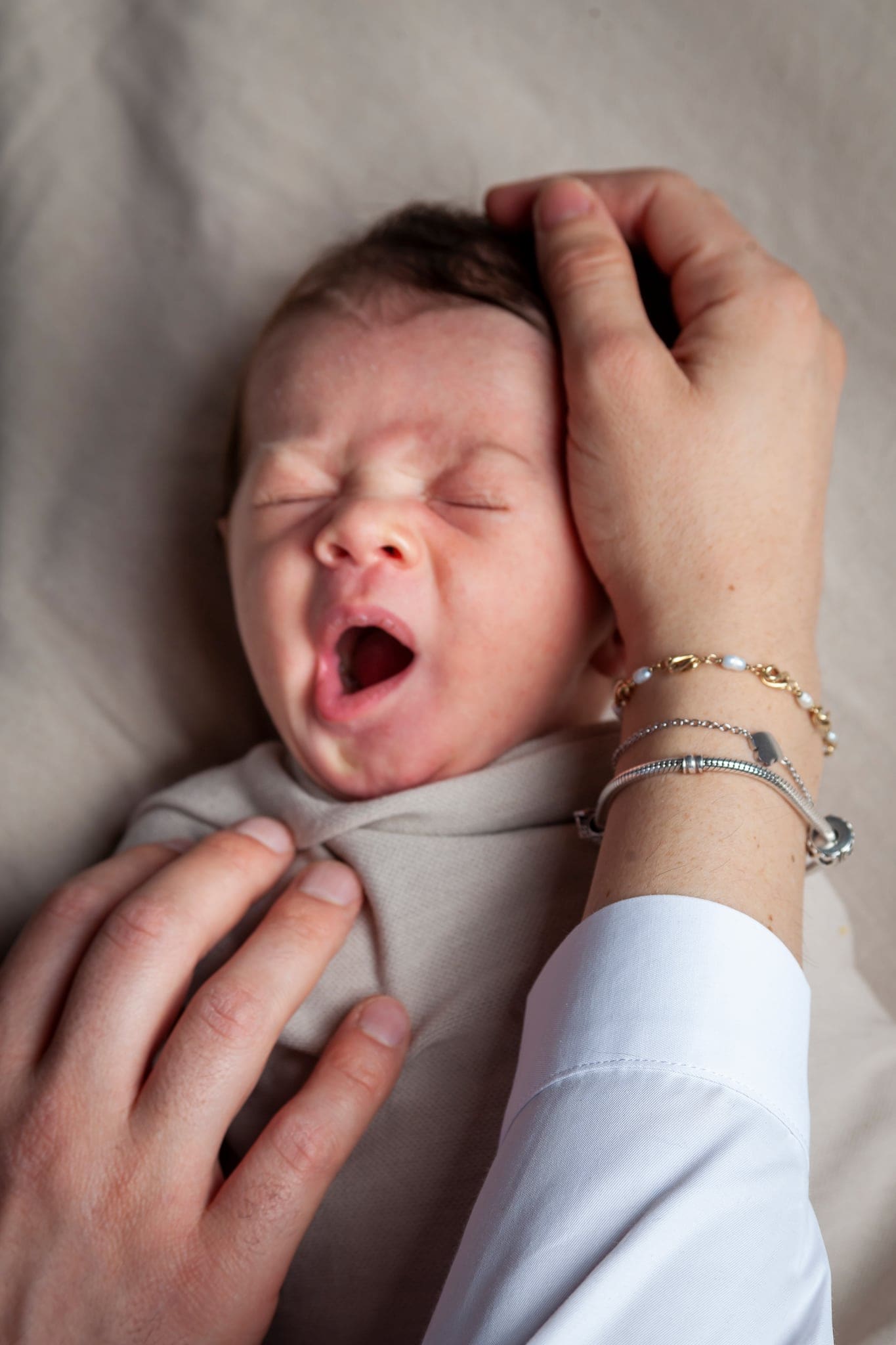 Neonato che sbadiglia a occhi chiusi con la mano della mamma sulla testa e un'altra sul fagotto beige.