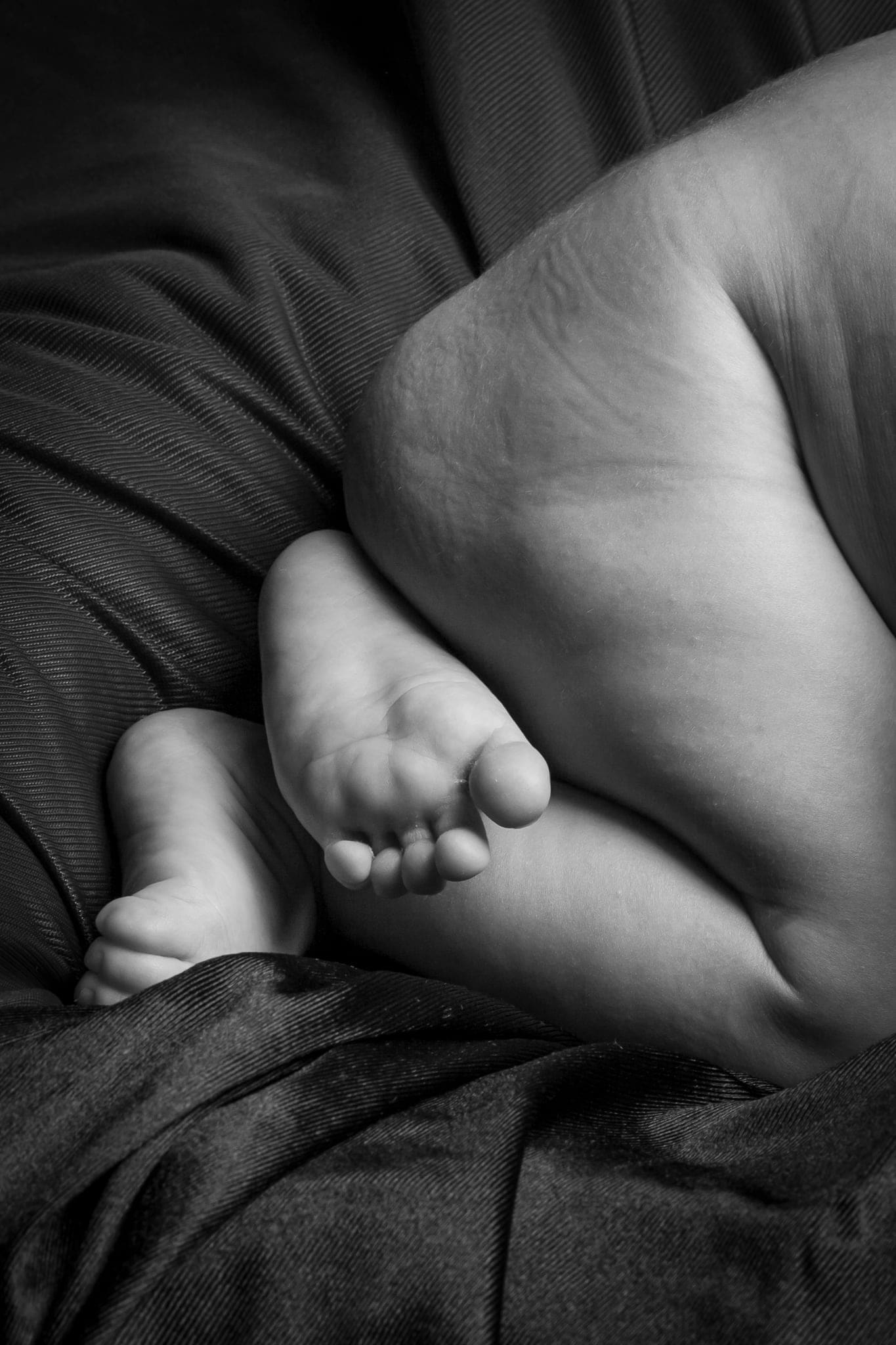Dettaglio dei piedini e delle gambine di un neonato raggomitolato, in bianco e nero su un tessuto lucido scuro.