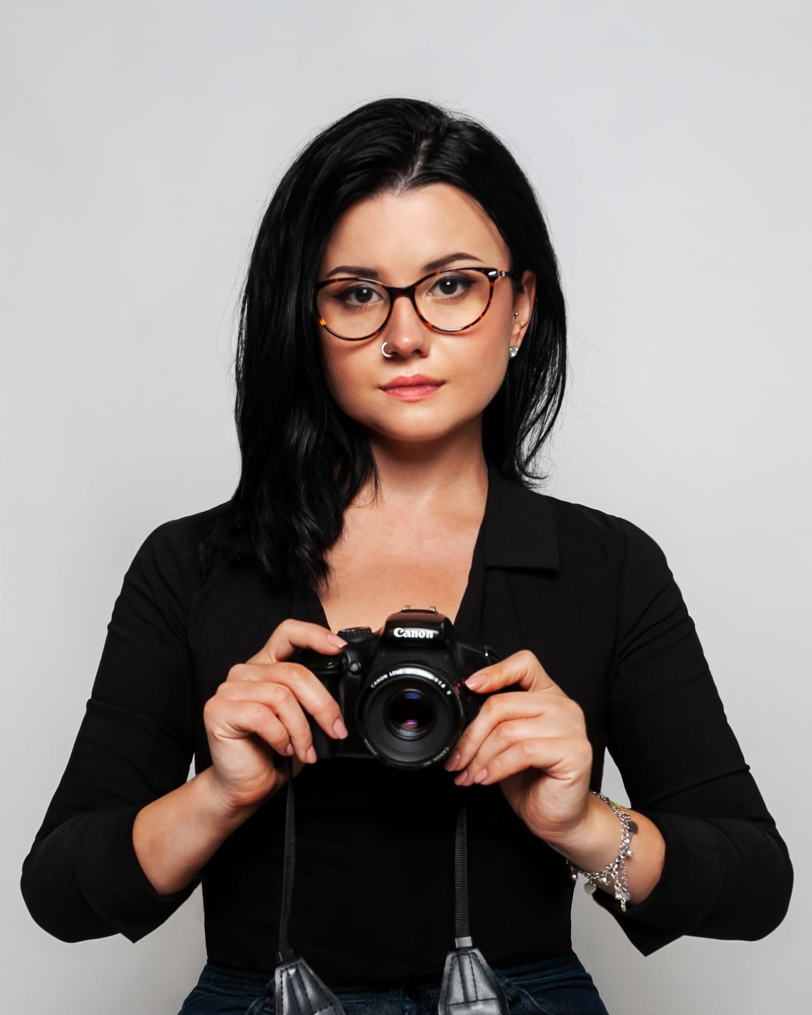 Viktoriya Hnenyuk, fotografa a Udine, con capelli scuri e occhiali, tiene la sua reflex davanti al petto su sfondo grigio chiaro